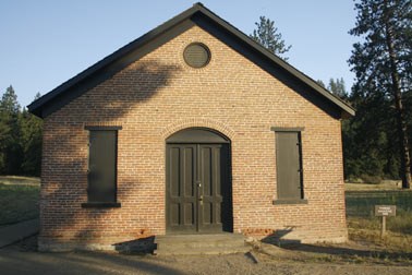 Brick building. Two windows, and a door between surrounding by ponderosa pines.