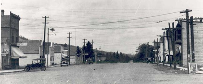 Cars on dirt road with wooden store fronts on either side.