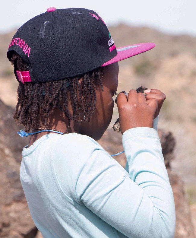 A child in a pink ball cap uses a magnifying glass to investigate a rock.