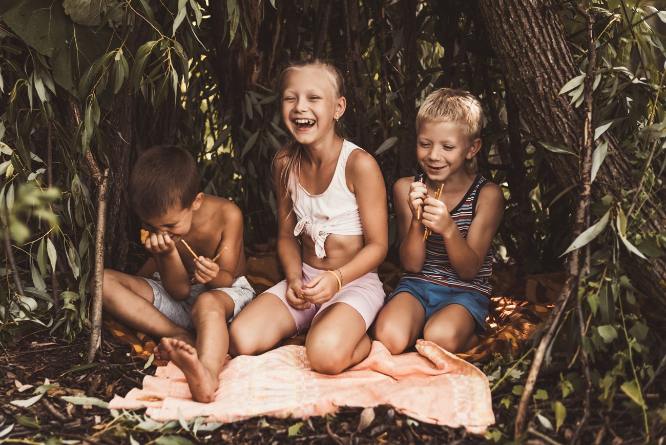 Three kids in a little hut made of branches, eating snacks and laughing hard.
