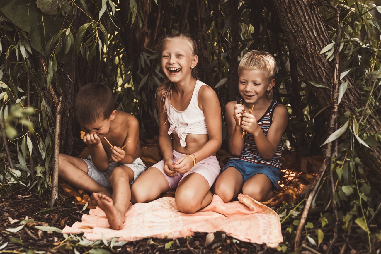 Kids playing and having fun Three kids in a little hut made of branches, eating snacks and laughing hard.