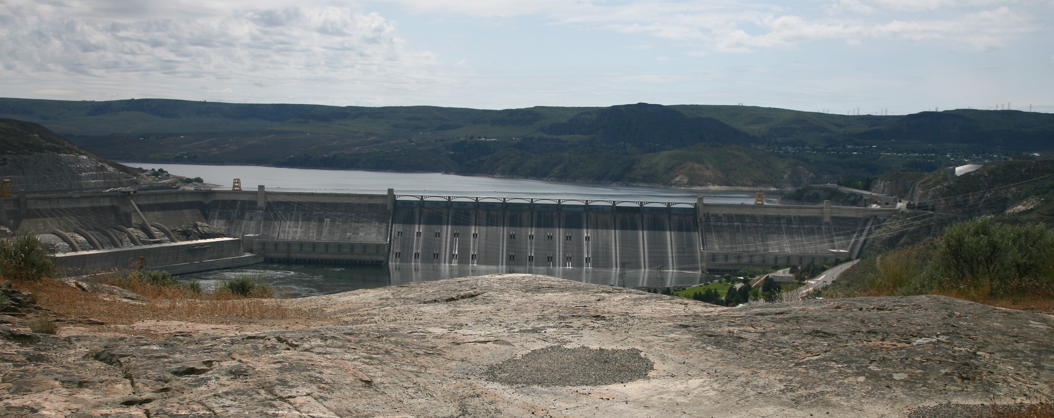 A view of Grand Coulee Dam from a cliff above it. The cliff is a smooth, light grey rock. Behind the concrete dam stretches a lake and rolling hills.