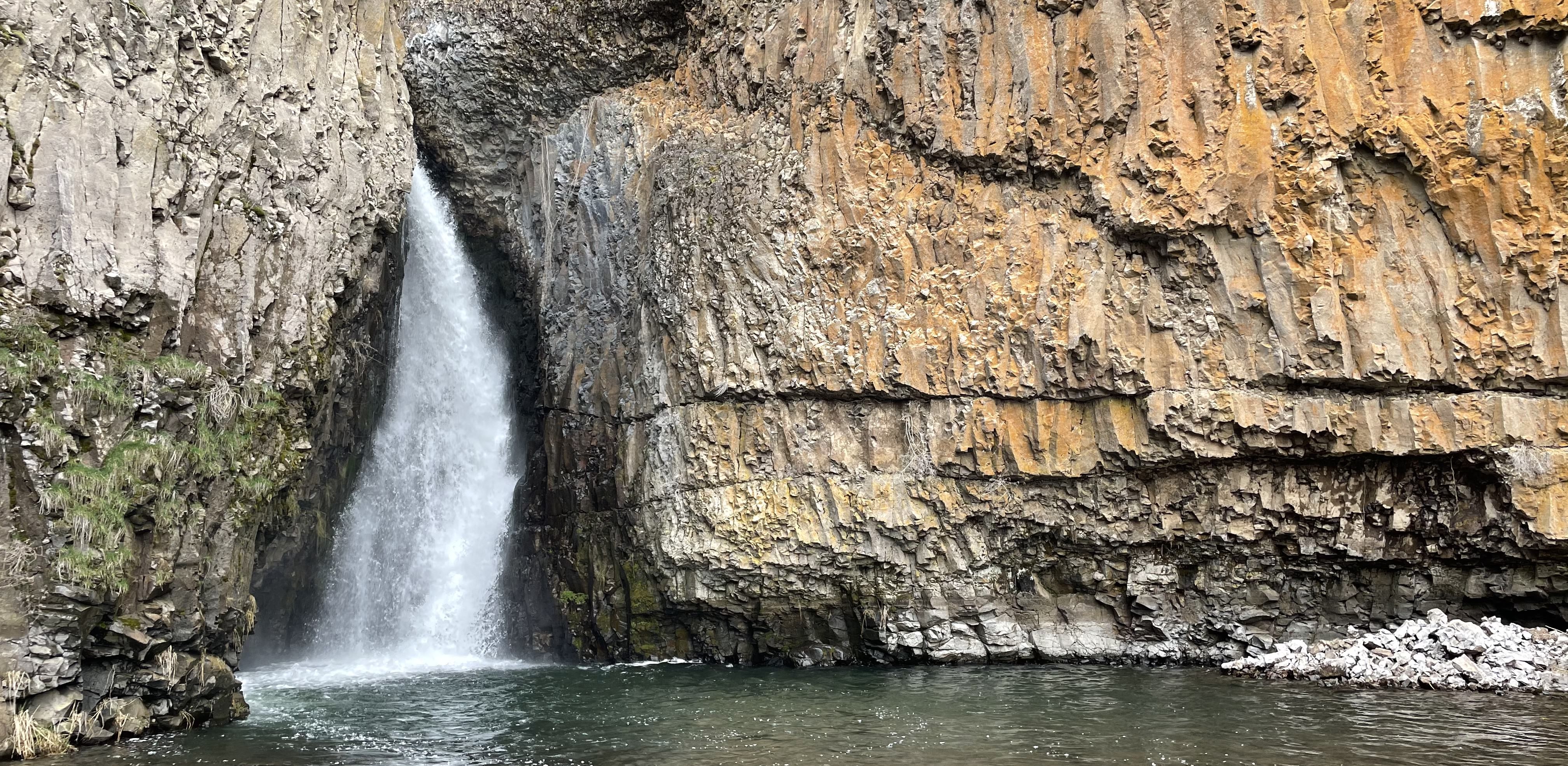 A 20ft waterfall pours from a small crack into a river. The rock surrounding the waterfall is dark with jointing and columns, covered in areas by orange lichen and green moss.