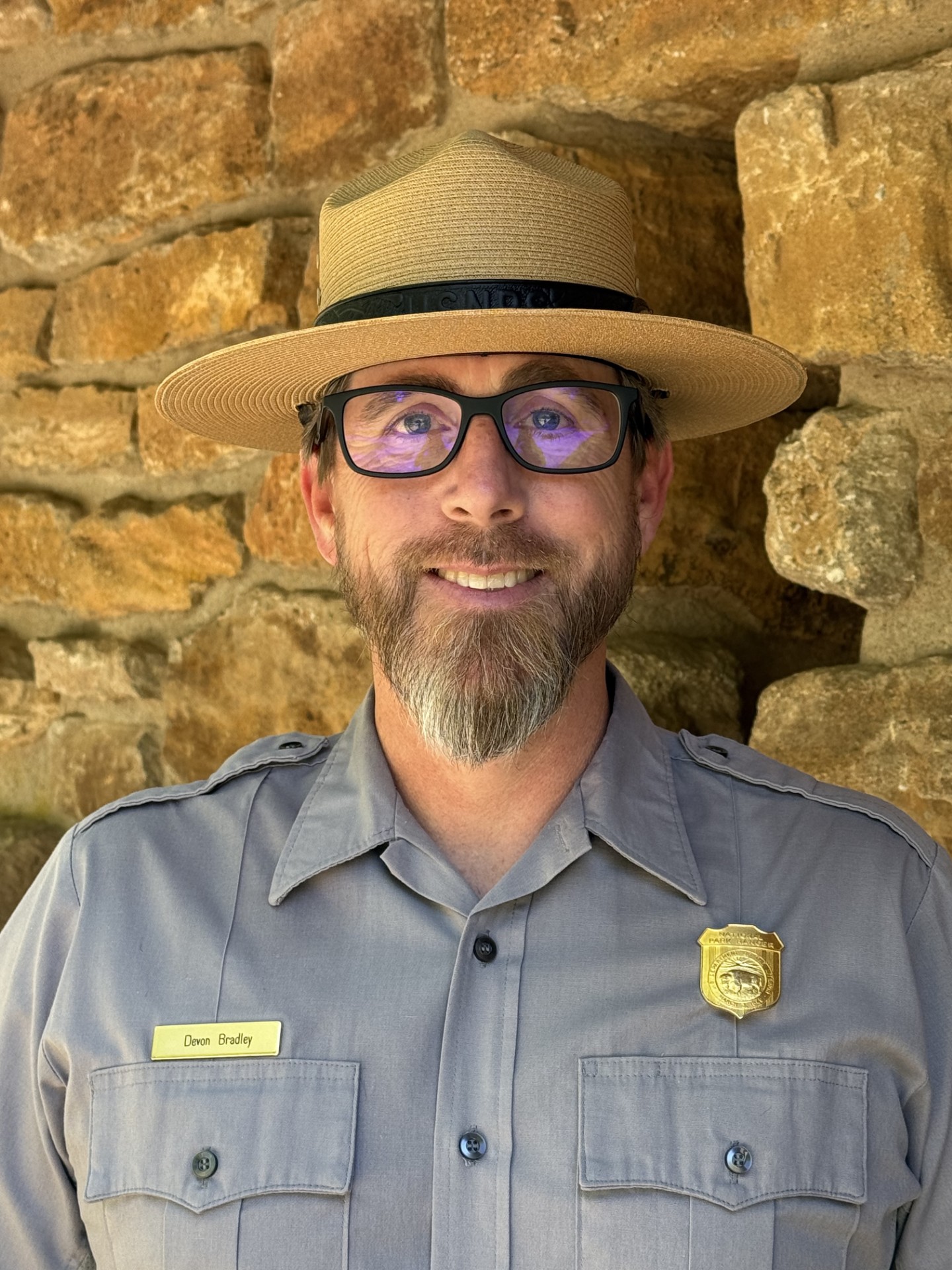 Portrait of a male park ranger with a wide brimmed straw hat and a grey buttondown shirt smiles for the camera.
