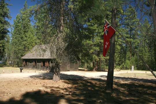St.Paul's Mission. One room, square log building set among pine trees. Hudson's Bay red flag in foreground.