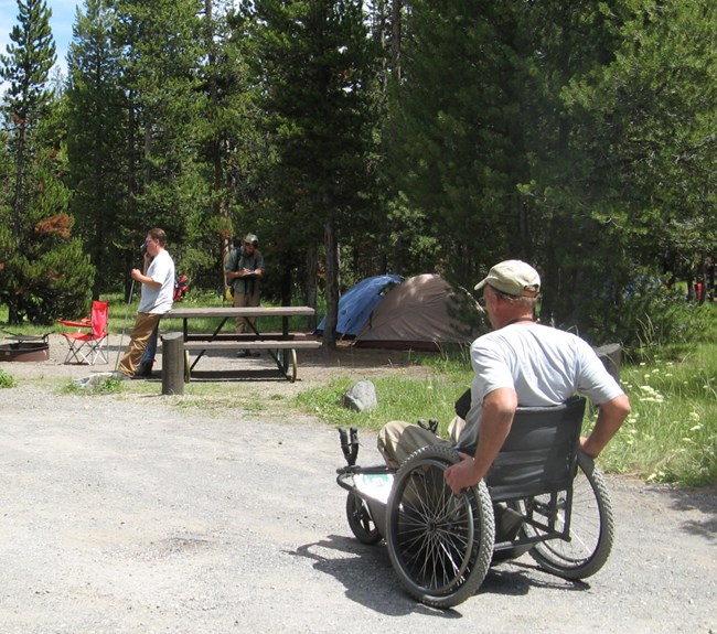 Man in manual wheelchair moves toward a campsite with two tents in it off of a dirt road.