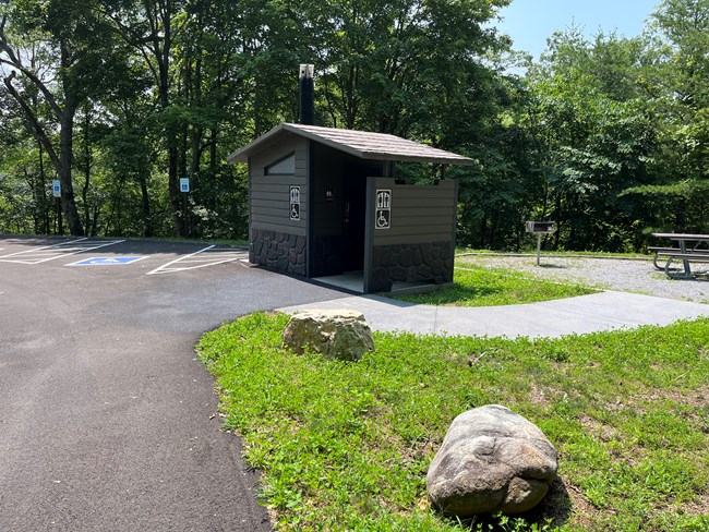 A brown restroom facility with a handicap sign on it, in addition to a restroom sign, beside a handicap accessible parking place and a picnic table.