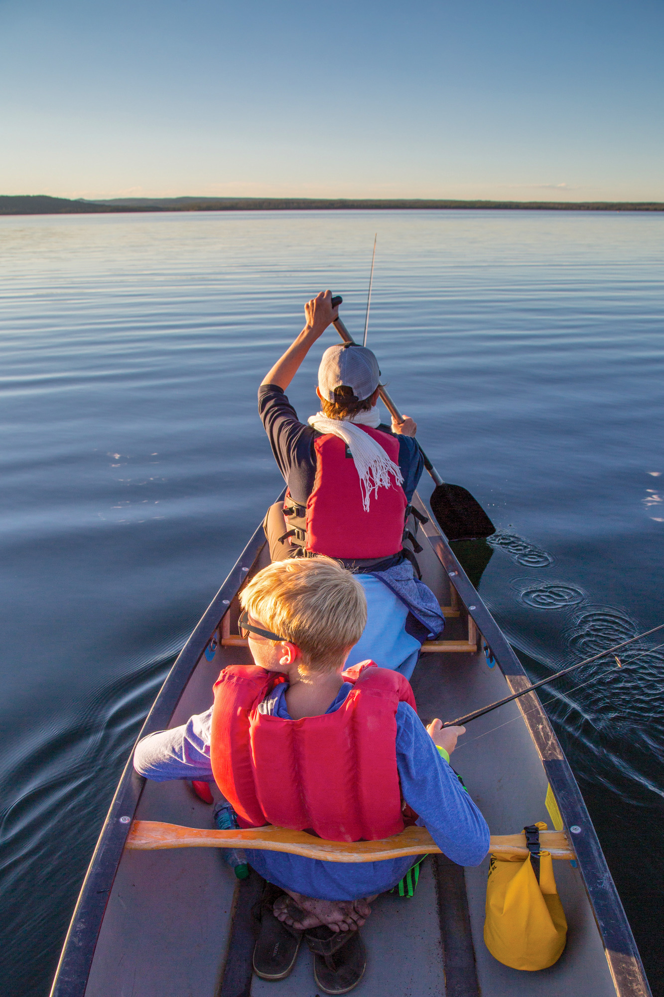 An adult and Child is in a canoe in calm water. The adult is paddling while the child holds a fishing pole