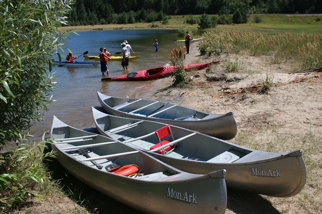 Three canoes rest on shore while children wearing life jackets sit in kayaks