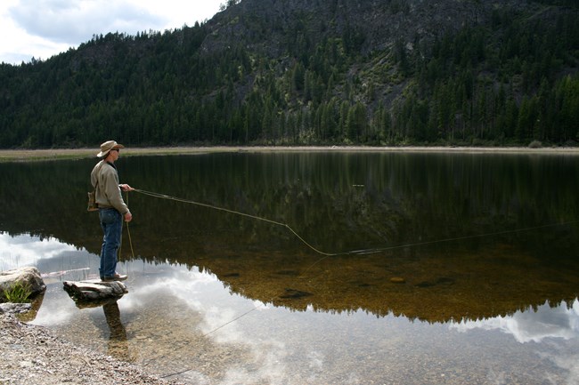 Fisherman stands on a small rock in water with fishing rod cast out into a lake.