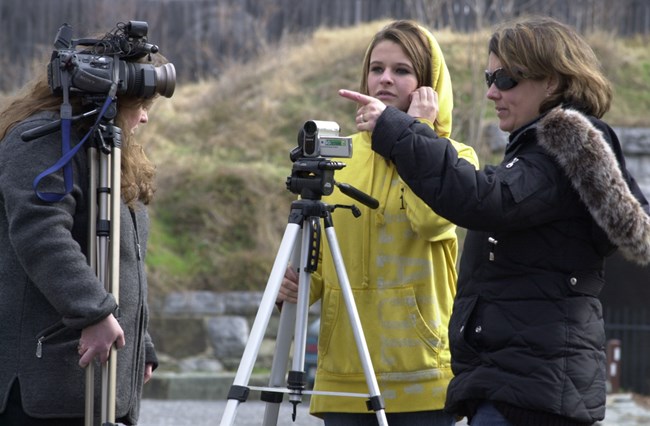 Two cameras are set up and a woman in a black jacket points into the distance.