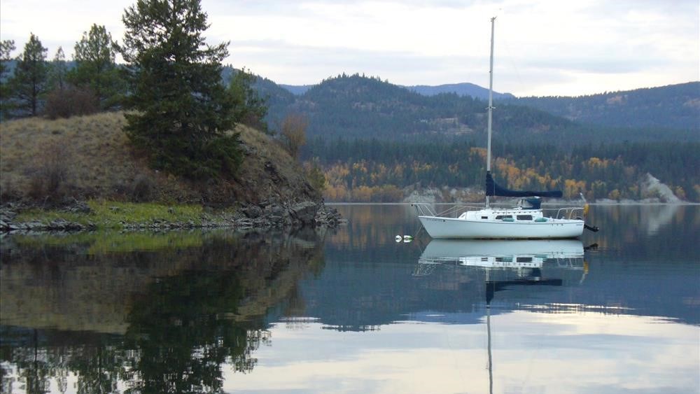 A sailboat sits on still water near a wooded shoreline. the still lake reflects the boat and shore