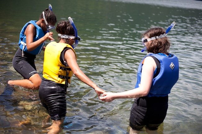 Three children wearing life jackets, face masks, and snorkels get ready for a swim in the lake.