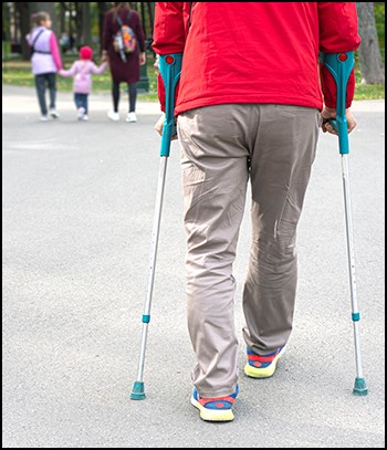 A person using crutches navigates a hardened walkway. Trees and another group are beyond.