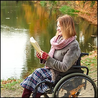 A woman dressed warmly on a fall day, sits in a wheelchair and reads a book next to a lake.
