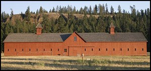 Long red wooden barn with many windows, black roof, and two chimneys. Ponderosa pine covers the hillside behind the building.