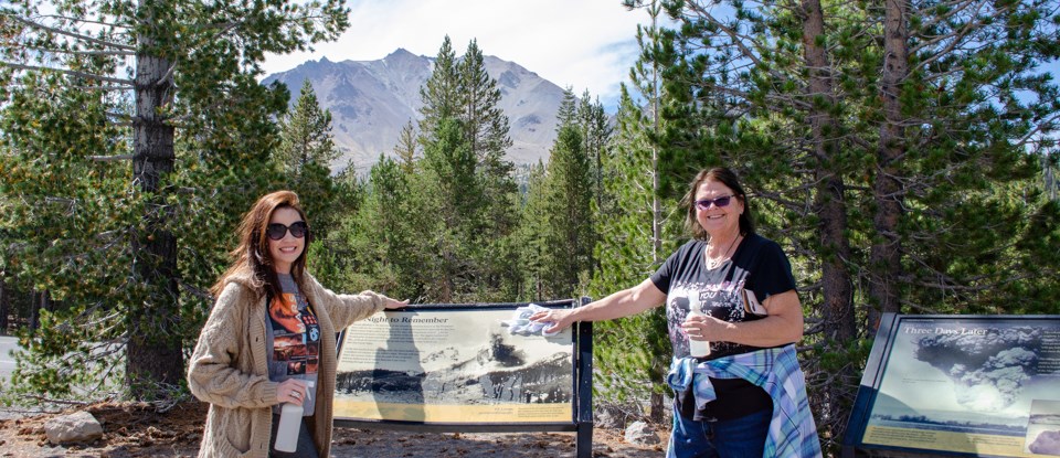 Two women hold cleaning supplies in front of a wayside