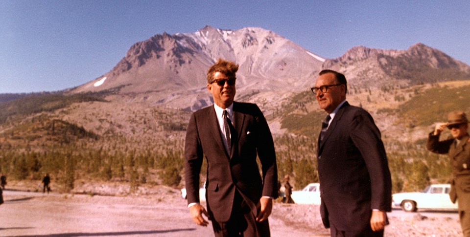Two men in black suits standing, backed by a bare volcanic peak. A ranger in a green uniform holds his hat down in the background at right.