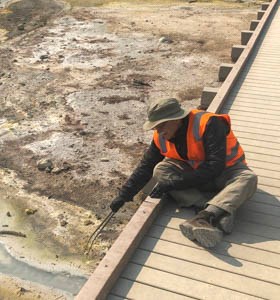 A seated man in an orange vests leans over a boardwalk to collect a sample with long tongs in a hydrothermal basin.