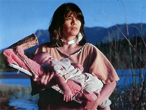 A woman in American Indian clothing holds a baby on a board backed by a lake and mountain.