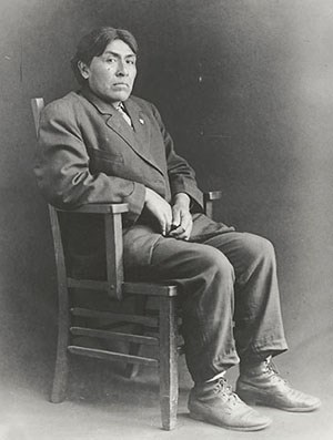 A man with short, dark hair in a dark jacket and pants sits in a chair.