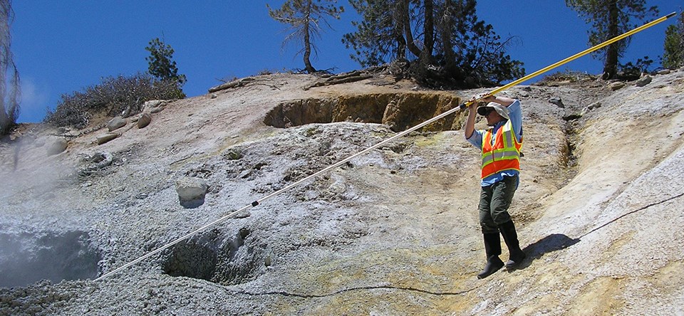 A man in an orange vests holds a long pole into a steaming mudpot