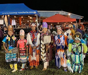 A group of kids and a woman stand in presentation attire at a POW WOW