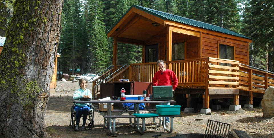 A man stands and a woman sits in a wheelchair at either end of a picnic table in front of a wooden cabin with an access ramp.