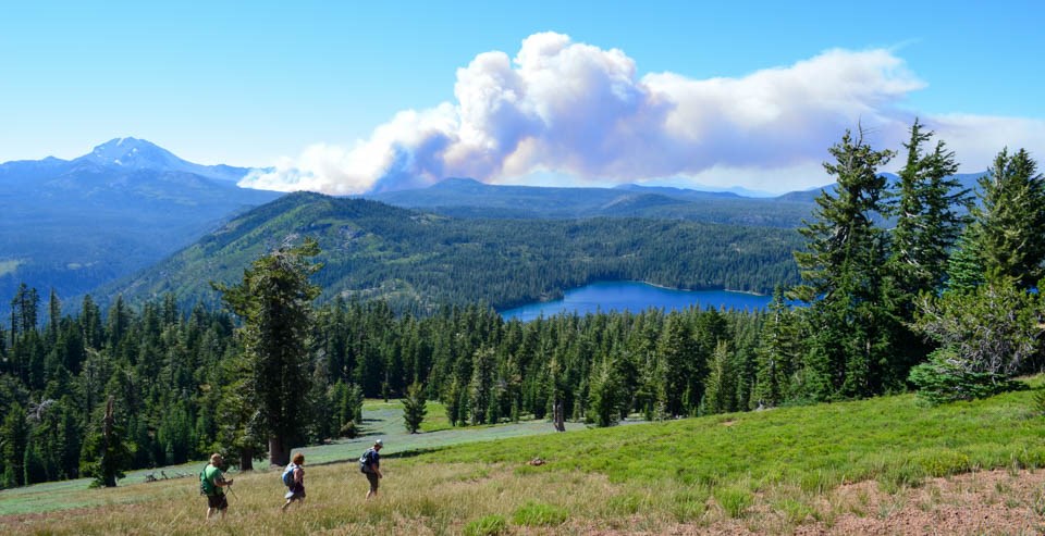 Three hikers cross a hillside above an alpine lake. A plume of smoke floats to the right from a wildfire in the distance.