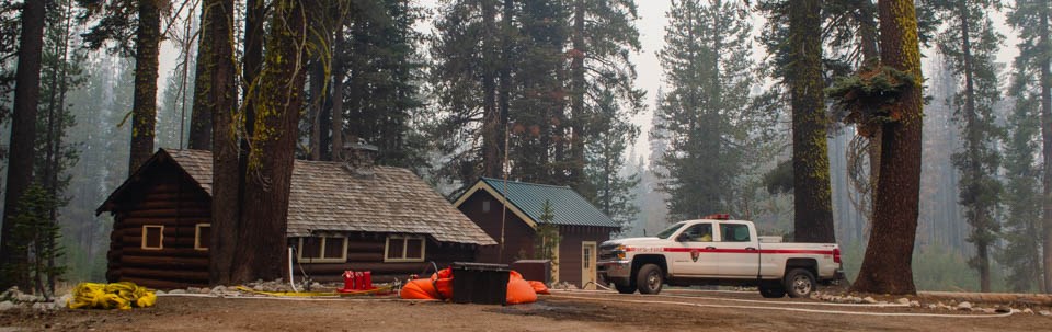 A white fire truck and structure protection equipment in front of two single-story, wooden  buildings amid conifer trees.