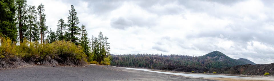 A dry lake bed lined by a lava flow, conifer trees, and brush.