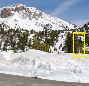 Multiple feet of snow along the edge of a plowed roadway backed by a volcanic peak. A tall snow course survey sign is highlighted in yellow.