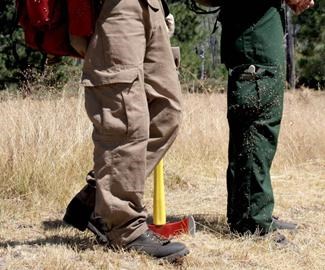 the legs of a firefighter wearing tan cargo pants holding an ax and a park ranger wearing green pants