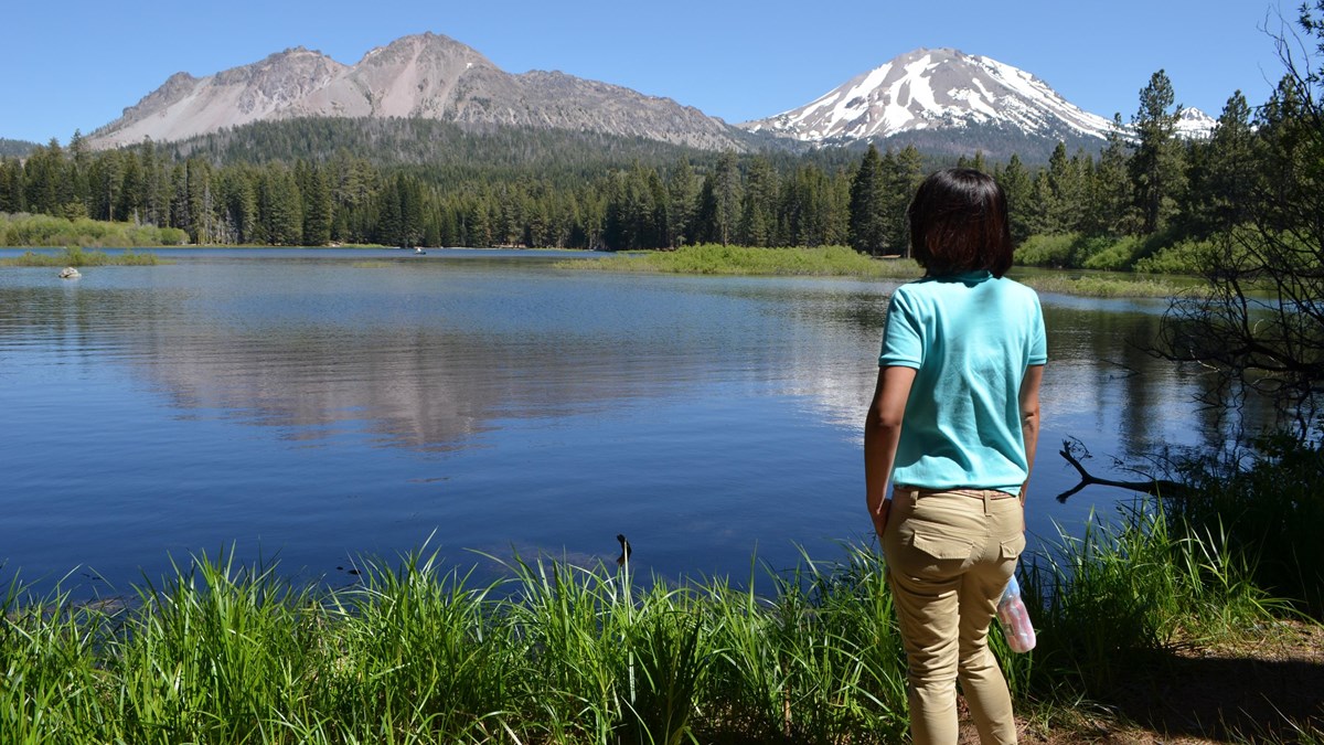 A person looks to the horizon in front of a lake with volcanoes in the background