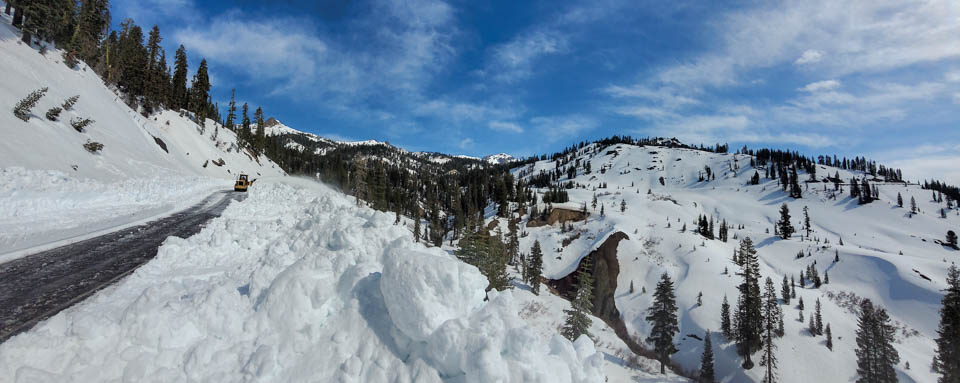 A panoramic photo of a snow-covered, mountain landscape. On the left side, a snow plow is barely visible on a newly plowed road.