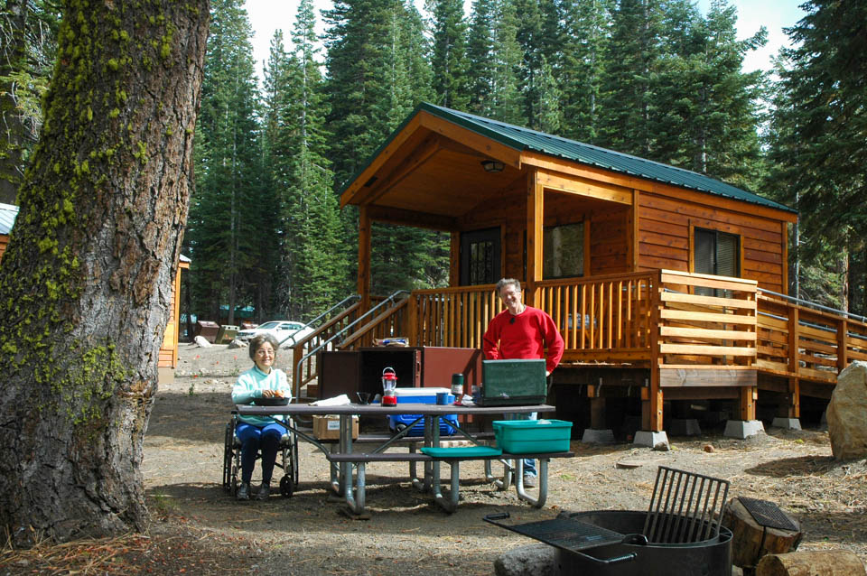 A man stands and a woman sits in a wheelchair at either end of a picnic table in front of a wooden cabin with an access ramp.