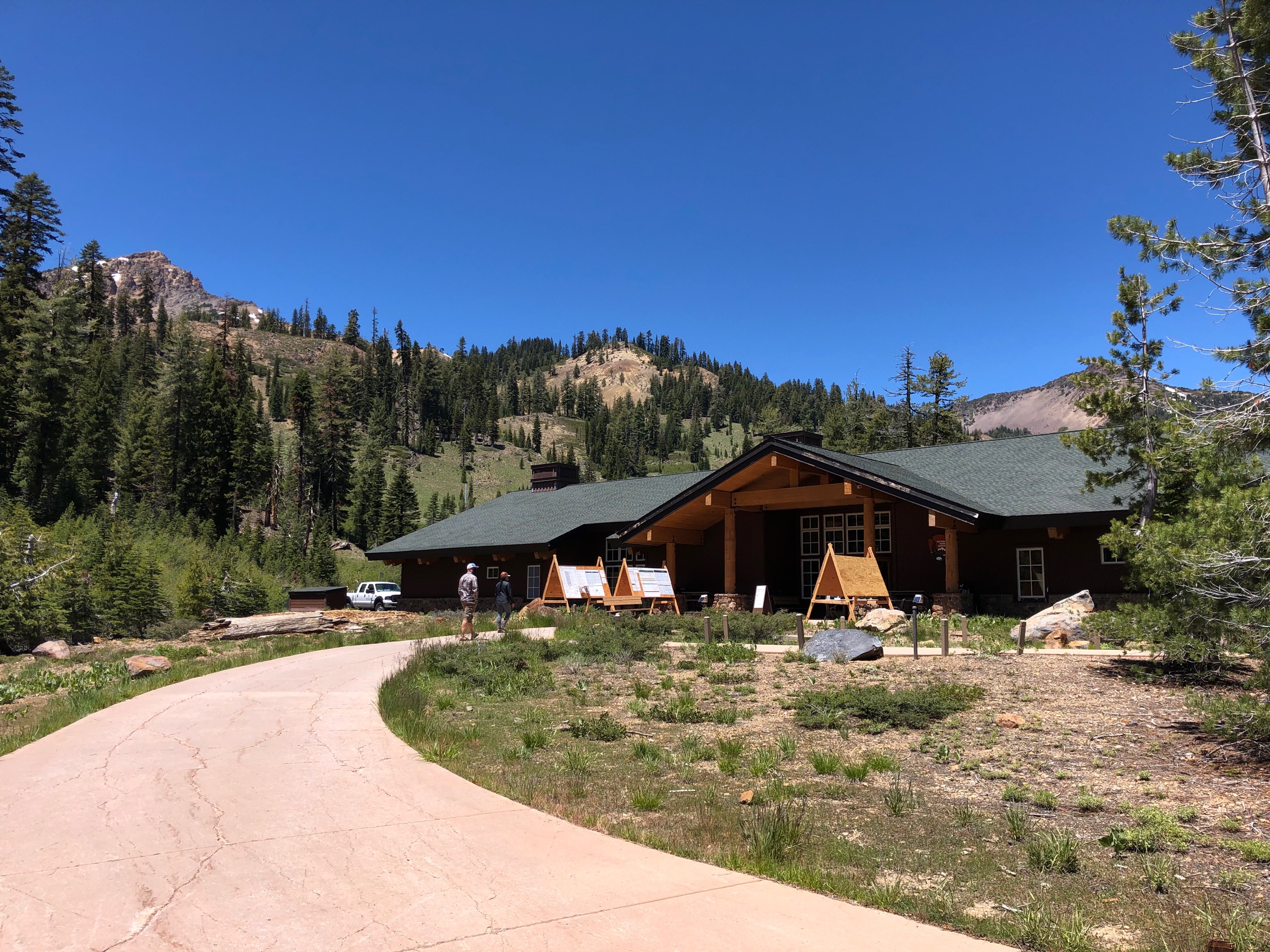 A tan walkway leads to a one-story wooden building backed by tree-covered mountains