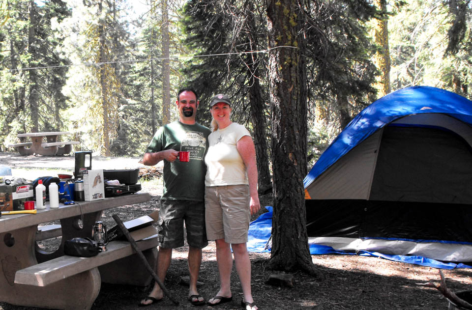 A man and a women stand between a picnic table and tent at a campsite.