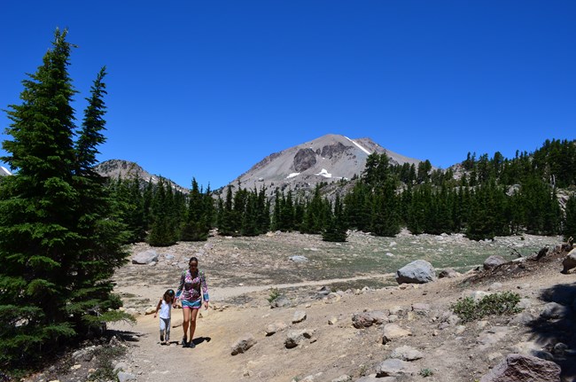 A woman and child walking on a dirt trail surrounded by pine trees and a tall mountain with patches of snow looms in the background.