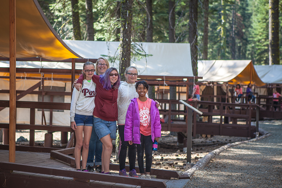 LAVO_DAL_3295_960W Five girls standing close pose for a photo on a wooden walkway outside of canvas tent at a youth camping facility