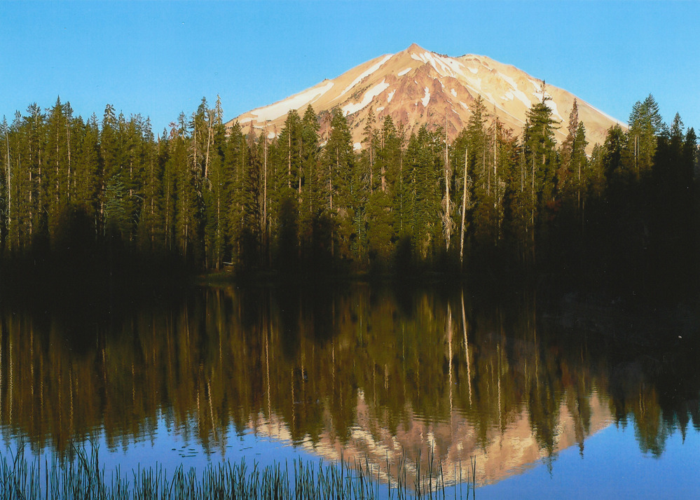 A photo of Lassen Peak, a tall volcanic peak nearly bare of snow, reflected in Summit Lake, a small lake with an exact upside down reflection of the mountain in the middle of the lake.