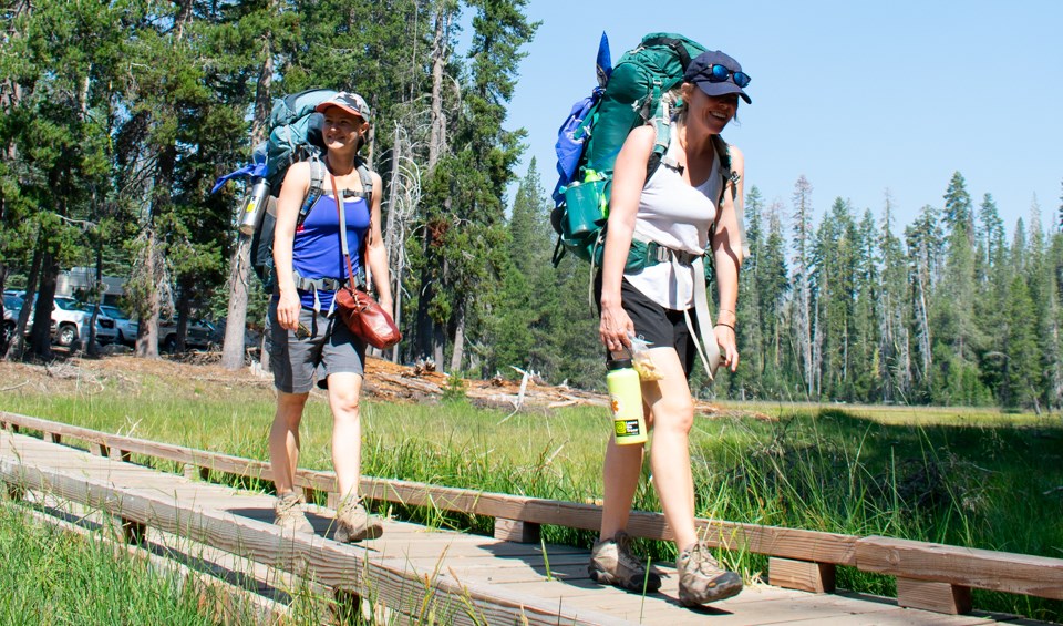 Two women wearing backpacks cross a wooden footbridge