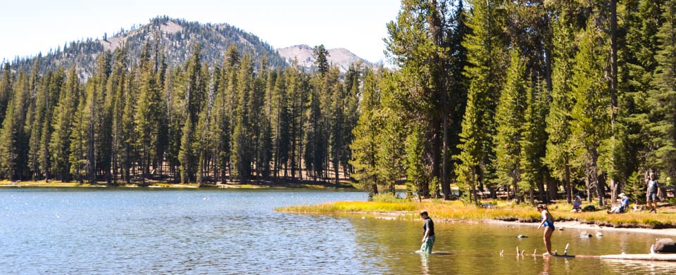 A boy and girl swimming in a mountain lake. Behind them, people site on a grassy shore.
