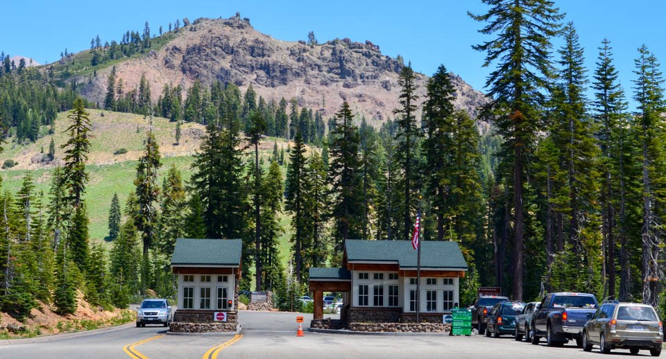 Two small buildings with a line of cars stopped to the right and a single car passing on the left.