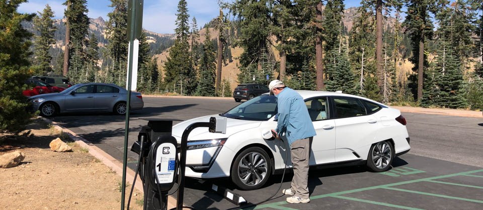 A man plugs an electric vehicle charger into a white vehicle in a parking area.