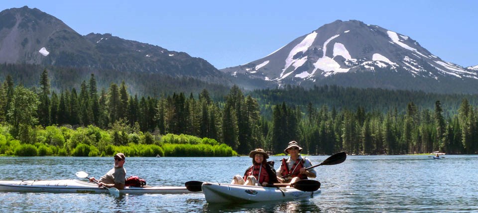 Three boaters in two kayaks pose for a photo on a lake backed by two large volcanic peaks.