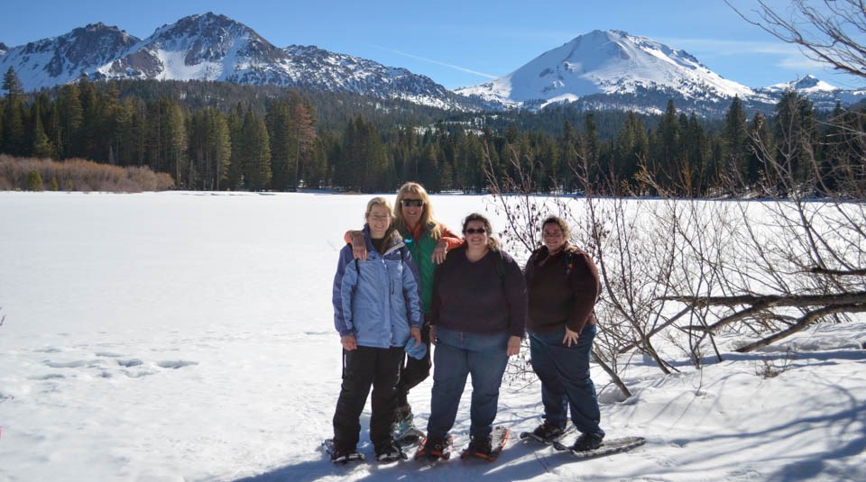 Four women on snowshoes at the edge of a frozen lake backed by large volcanic peaks.