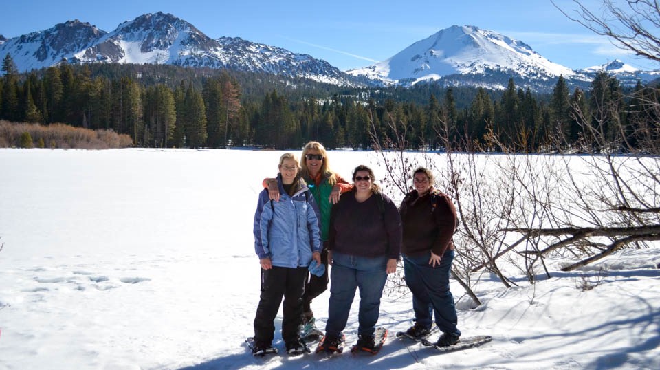 Four women on snowshoes pose for a photo in front of a front lake backed by volcanic peaks.