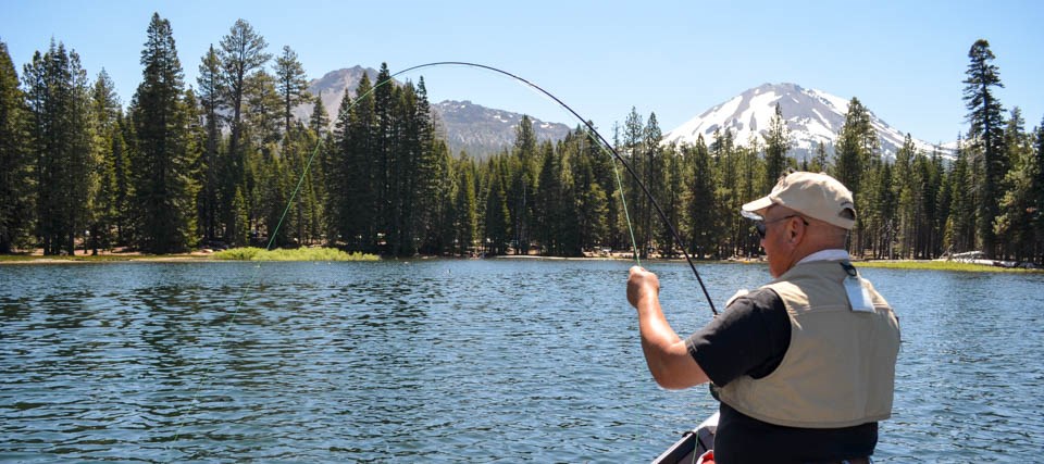 A man in a beige fishing vest in a boat holds a rounded fishing line. A snow-capped mountain rises above green conifers in the midground.