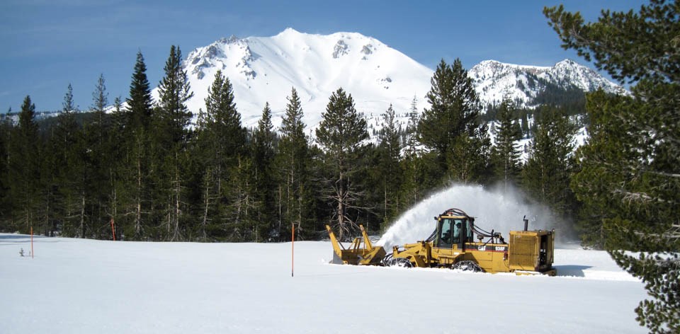A yellow snowplow clears snow backed by a snow-covered mountain
